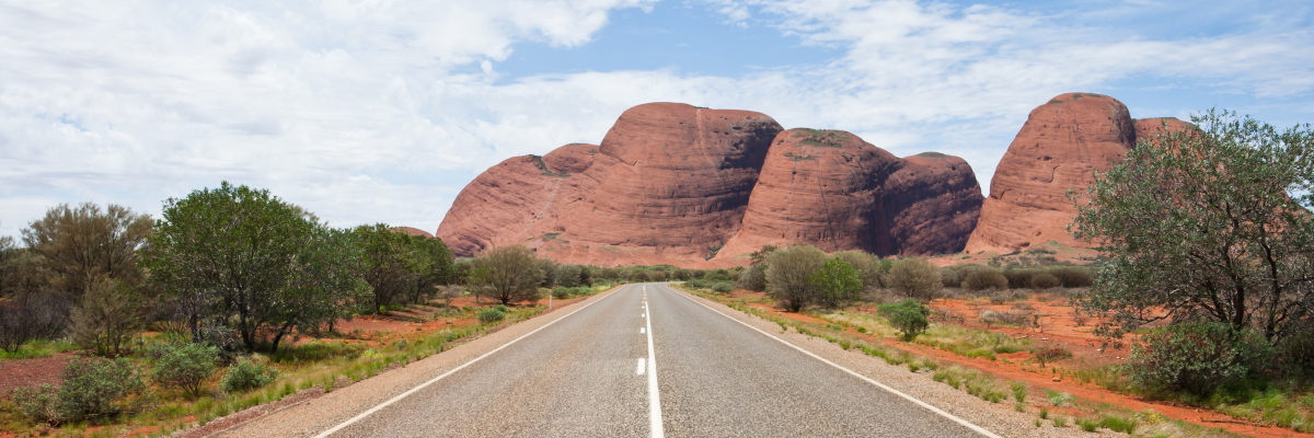 Photograph of a straight road cutting through the red sand of the outback, leading towards boulder-like rock formations lay ahead.